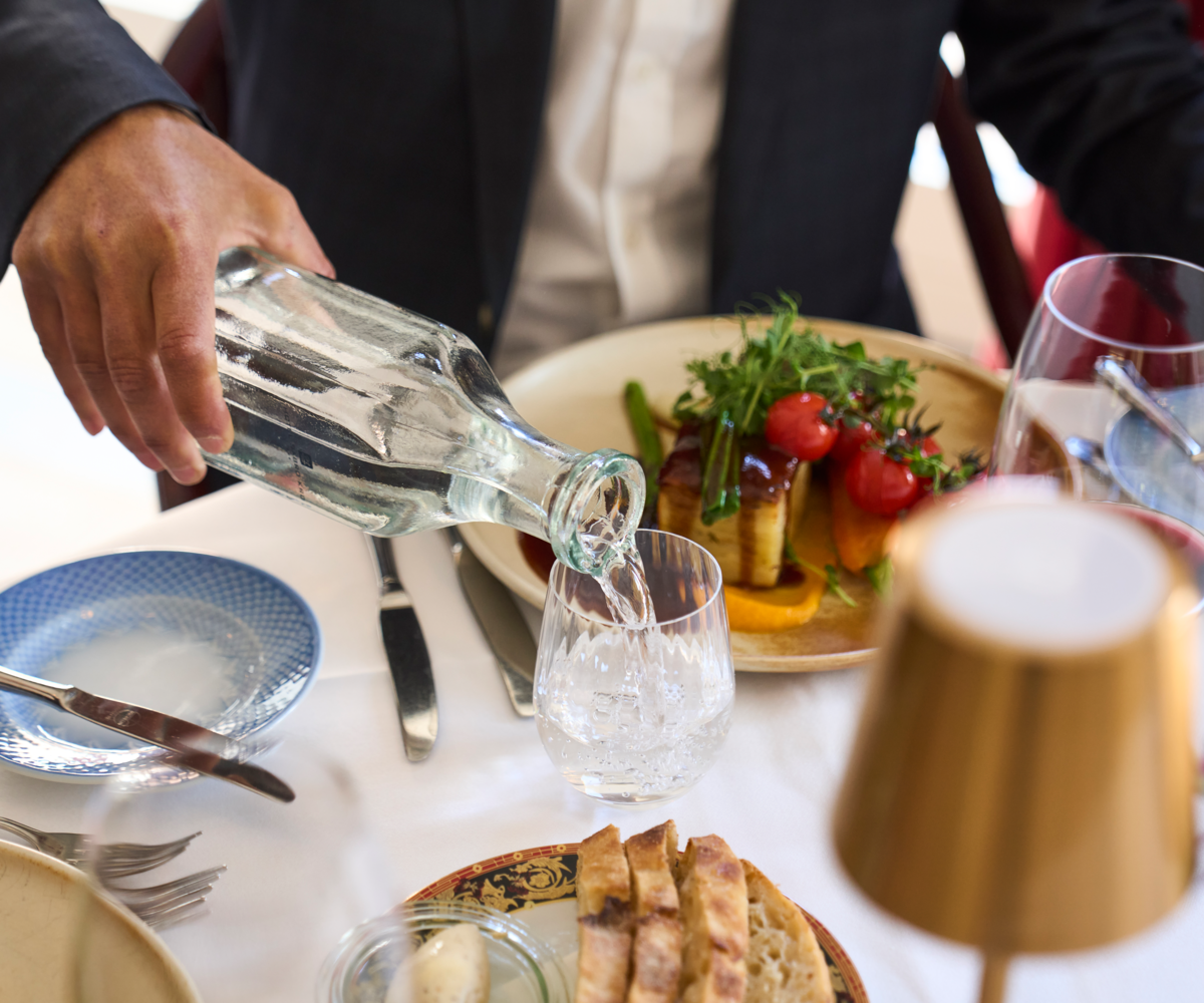 Vand hældes op i et glas ved siden af en smukt anrettet frokosttallerken med bagt selleri, grillede tomater og sauce – et elegant frokostbord på Hotel Sabro Kro.