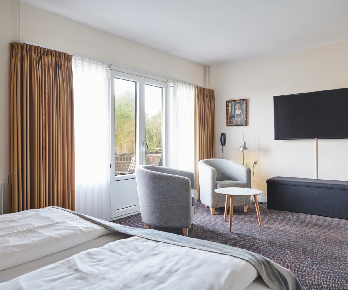 Lyst og rummeligt hotelværelse med dobbeltseng, to lænestole og tv, samt adgang til altan gennem dobbeltdøre med gardiner.