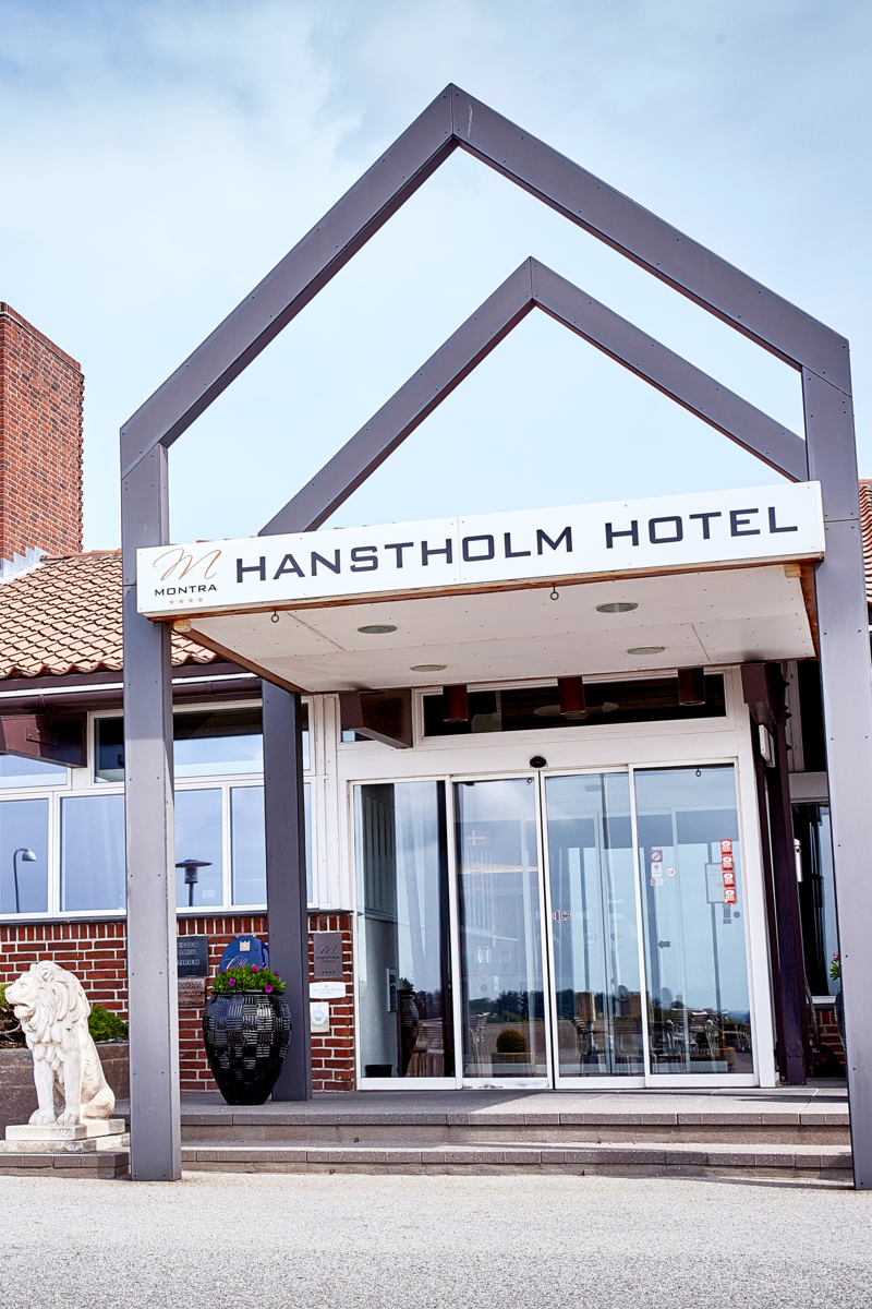 Facadeindgang til Montra Hotel Hanstholm med glidedøre, moderne tagkonstruktion og skilt med hotellets navn – flankeret af blomsterkrukke og stenskulptur af en løve.