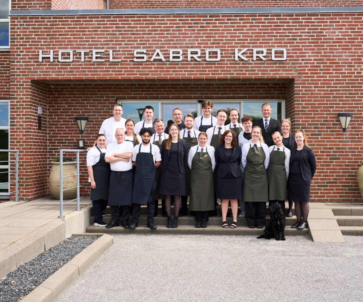 Gruppebillede af medarbejderne på Montra Hotel Sabro Kro, der står foran hotellets røde murstensfacade i deres uniformer. Med stiindgang på venstre side og store krukker med forårsblomster på højre side.
